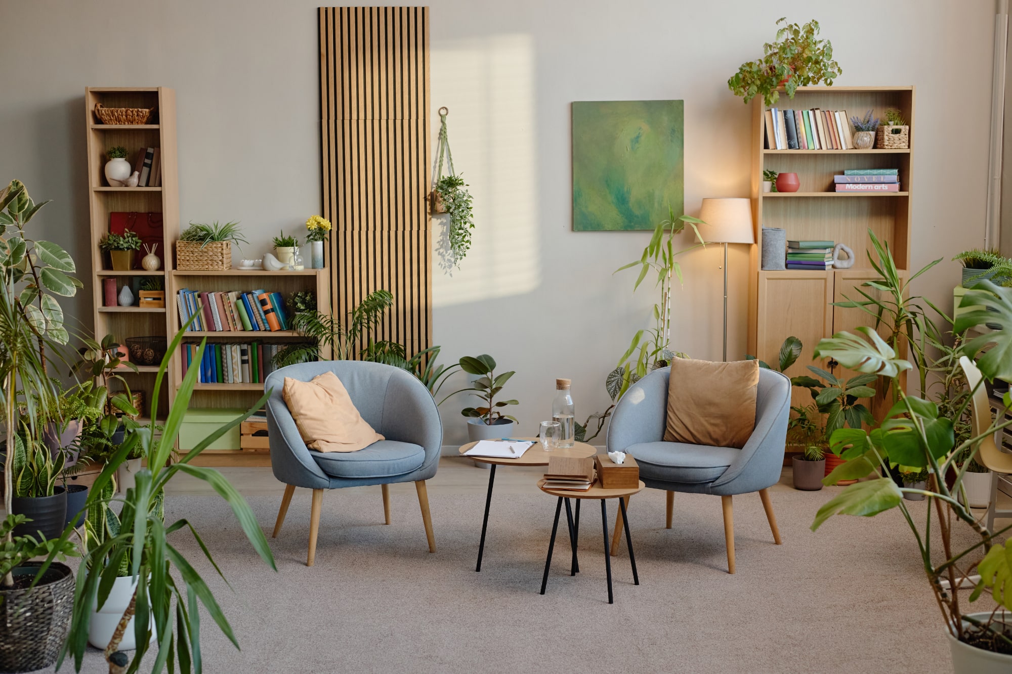 Deux fauteuils modernes, munis de coussins, font face à une petite table dans un salon rempli d'étagères, de plantes en pot, de livres et d'œuvres d'art accrochées aux murs, le tout baigné de lumière naturelle.