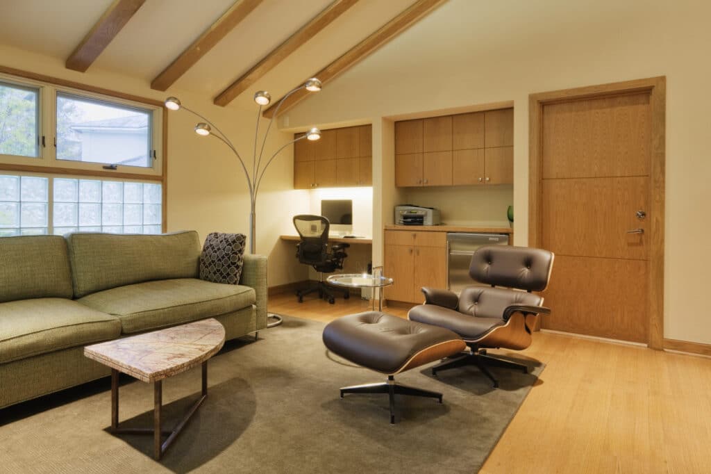 Salon moderne avec un canapé vert, un fauteuil Eames, un lampadaire en forme d'arc, un bureau encastré avec une chaise de bureau et des meubles de rangement en bois, le tout devant un mur beige.