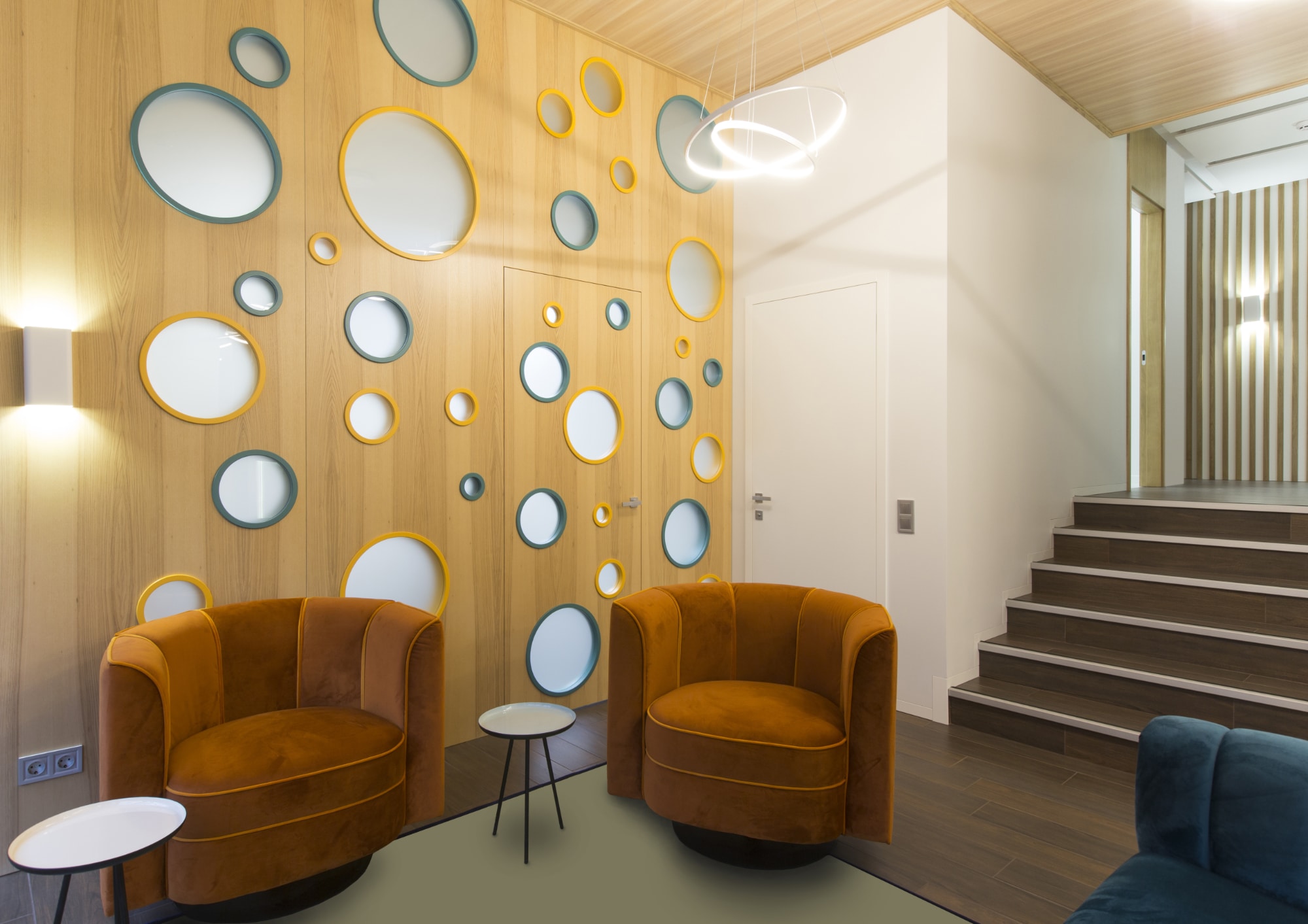 Salon moderne avec deux fauteuils orange, des tables d'appoint rondes et un mur en bois orné de miroirs circulaires de différentes tailles. Un escalier mène à un espace surélevé situé à droite. Des luminaires géométriques sont suspendus au plafond.