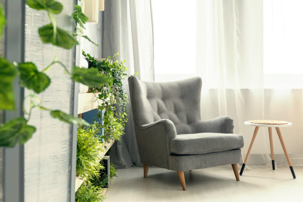 Un fauteuil gris est posé à côté d'une petite table d'appoint ronde, près d'une fenêtre lumineuse, tandis que des plantes vertes agrémentent l'espace.