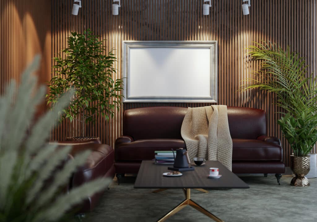 Salon moderne avec des canapés en cuir marron, des plantes vertes, une table basse noire et un tableau vierge encadré accroché à un mur en lattes de bois, le tout baigné d'une lumière chaleureuse grâce à des spots.