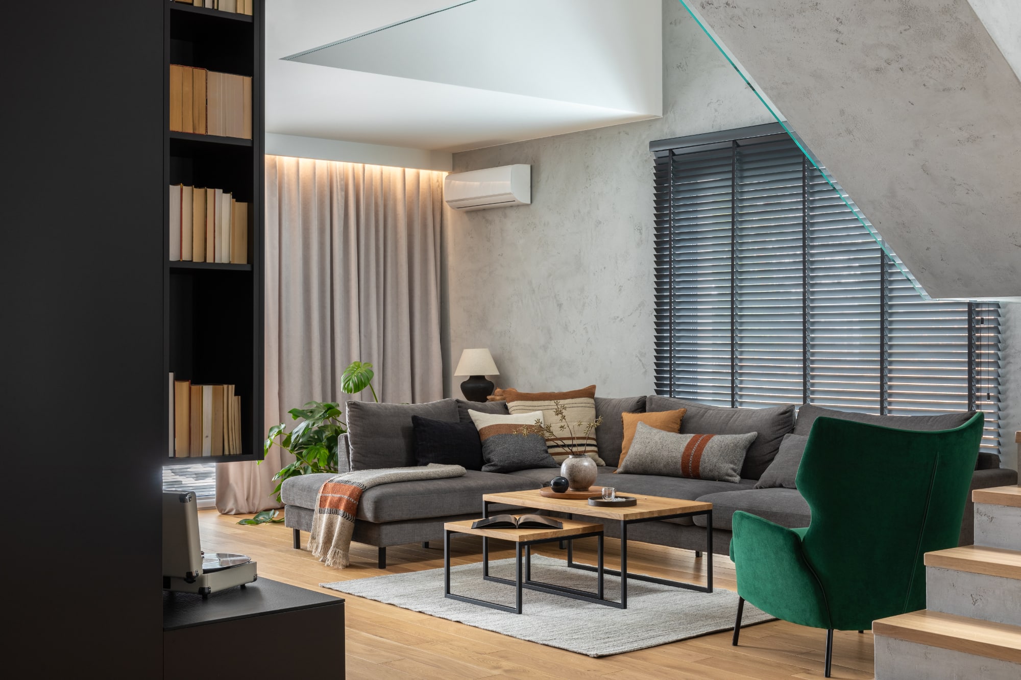 Salon moderne avec un canapé modulable gris, un fauteuil vert, des tables basses, des bibliothèques, un parquet et une fenêtre équipée de stores noirs. Décoration aux tons neutres et plante en pot dans un coin.