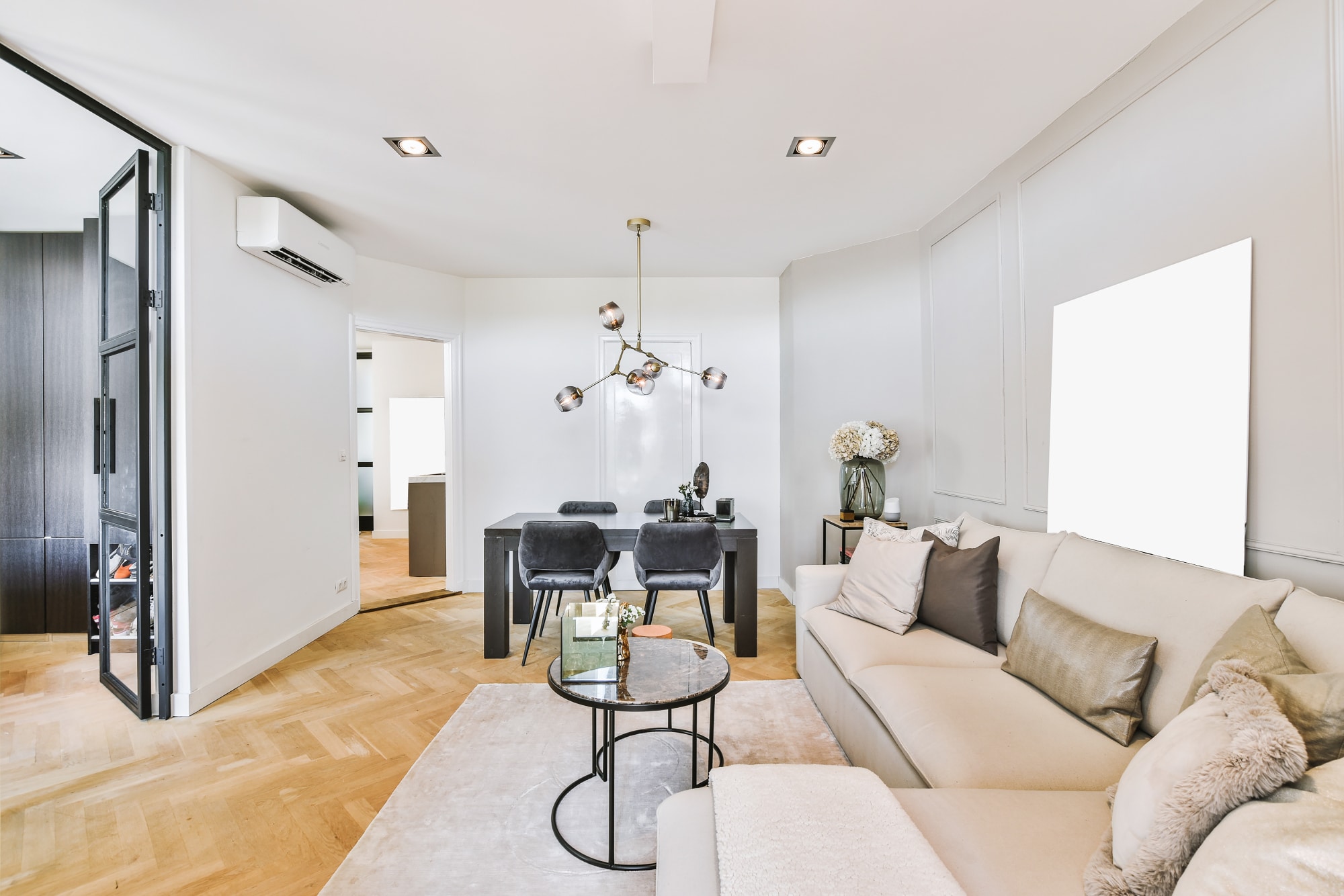 Salon moderne avec un canapé beige, des tables basses rondes, un coin repas avec des chaises noires, du parquet et une décoration minimaliste sous un éclairage lumineux.