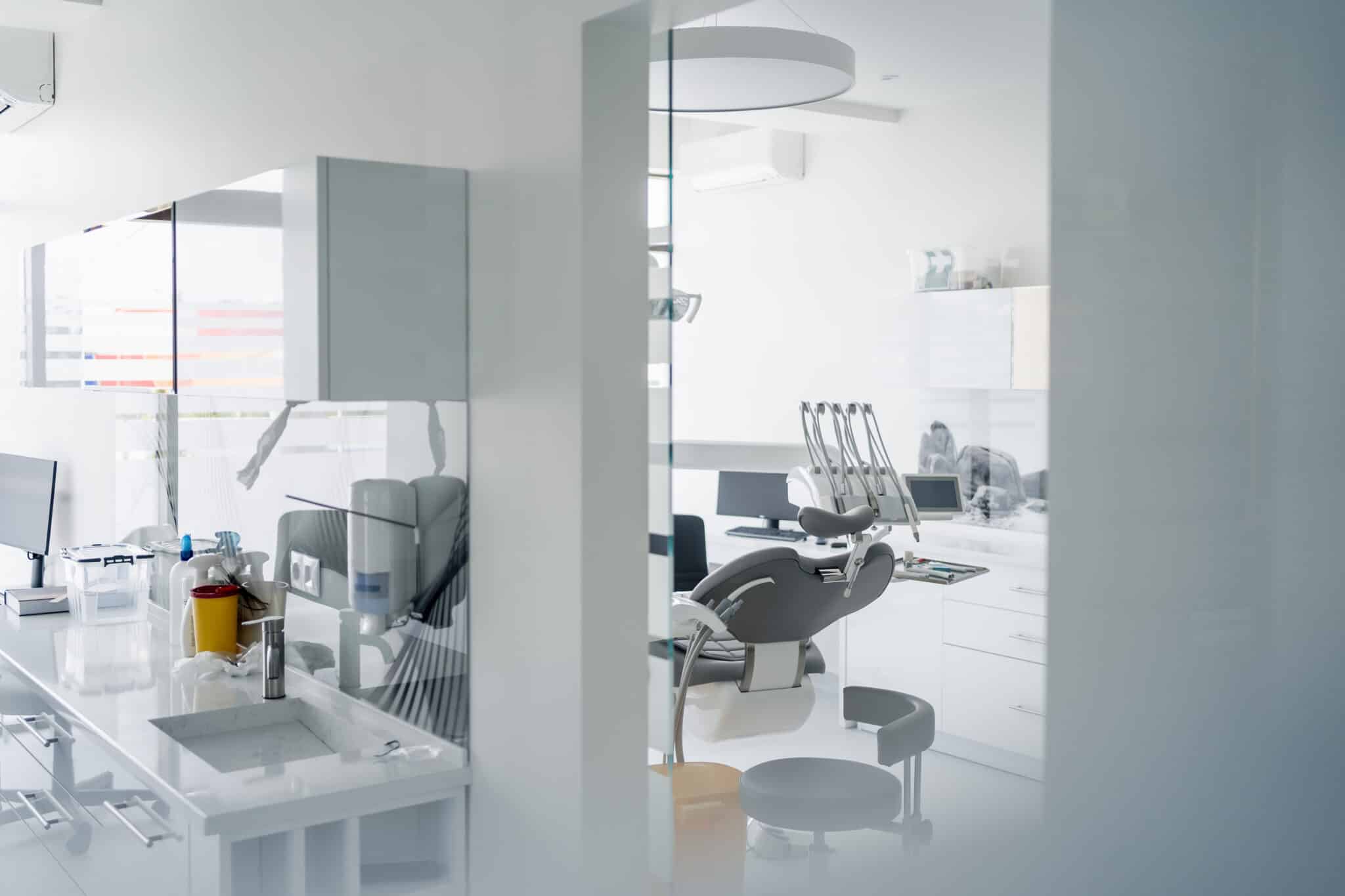 Cabinet dentaire moderne avec un fauteuil dentaire inoccupé, des armoires blanches, des écrans d'ordinateur et divers instruments dentaires posés sur les plans de travail, vu à travers une cloison vitrée.