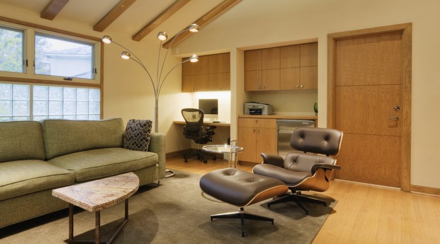 Salon moderne avec un canapé vert, un fauteuil Eames, un lampadaire en forme d'arc, un bureau encastré avec une chaise de bureau et des meubles de rangement en bois, le tout devant un mur beige.