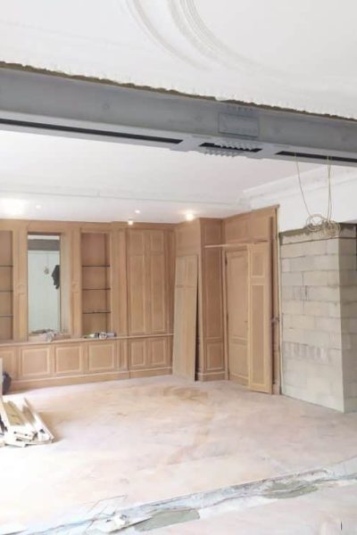 Une pièce en cours de rénovation avec une poutre en acier apparente, des murs non finis et des armoires en bois encastrées le long du mur du fond. Des matériaux de construction sont éparpillés sur le sol.
