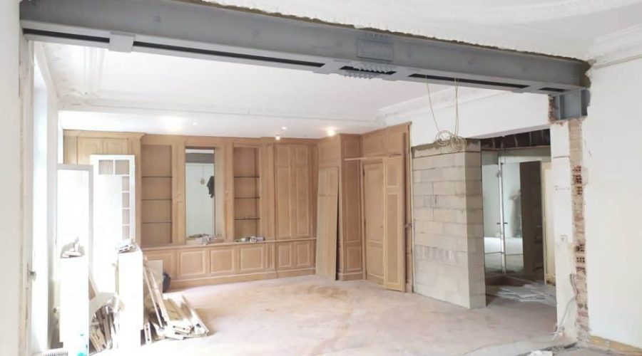 Une pièce en cours de rénovation avec une poutre en acier apparente, des murs non finis et des armoires en bois encastrées le long du mur du fond. Des matériaux de construction sont éparpillés sur le sol.