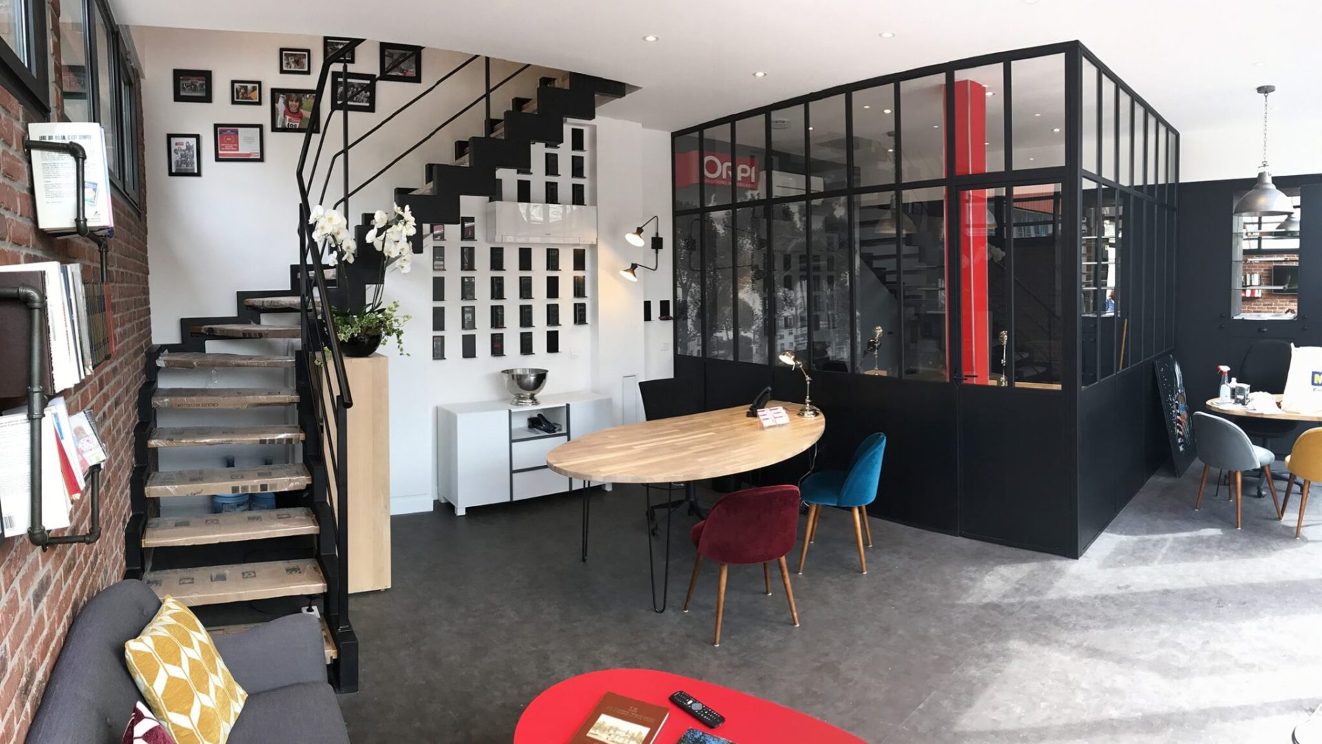 Espace de bureau moderne avec une salle de réunion vitrée, des escaliers avec des photos murales, une table ronde en bois avec des chaises colorées et divers objets de décoration.