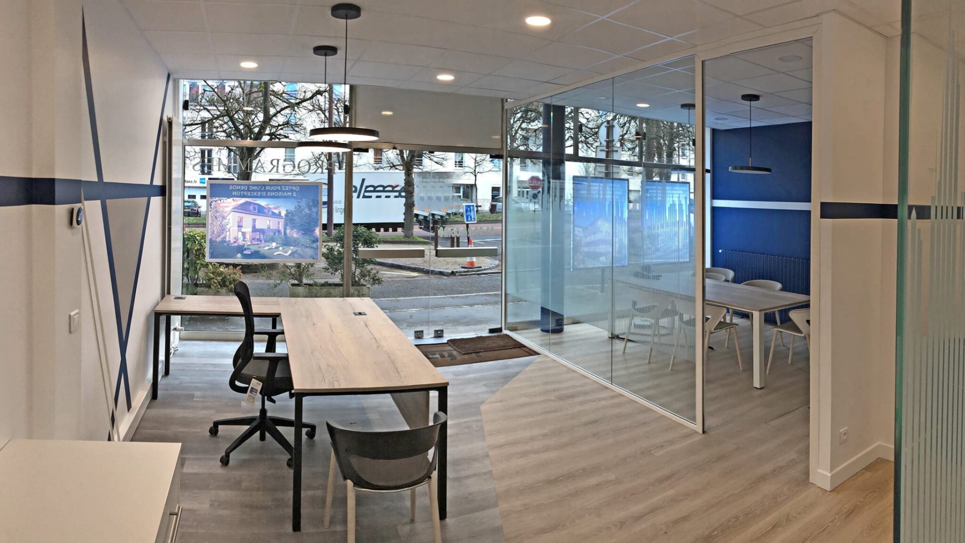Bureau moderne avec de grandes fenêtres, deux bureaux et des chaises au premier plan, et une salle de réunion vitrée avec une table et des chaises à l'arrière-plan.