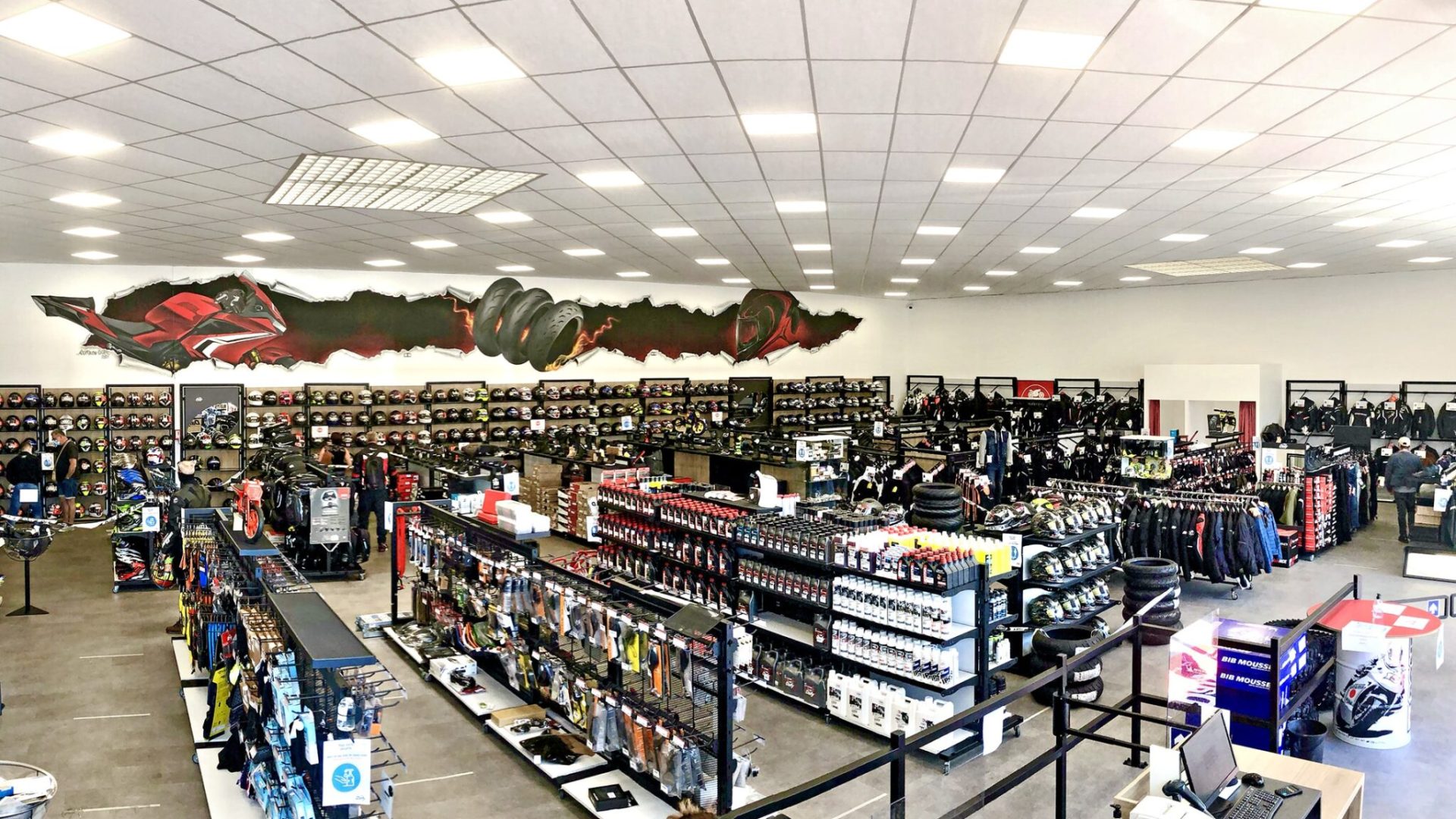 Vue panoramique d'un magasin d'équipement et d'accessoires pour motocycles présentant des casques, des vestes, des gants, des pièces détachées et des vêtements sur des étagères et des rayonnages sous un éclairage zénithal lumineux.