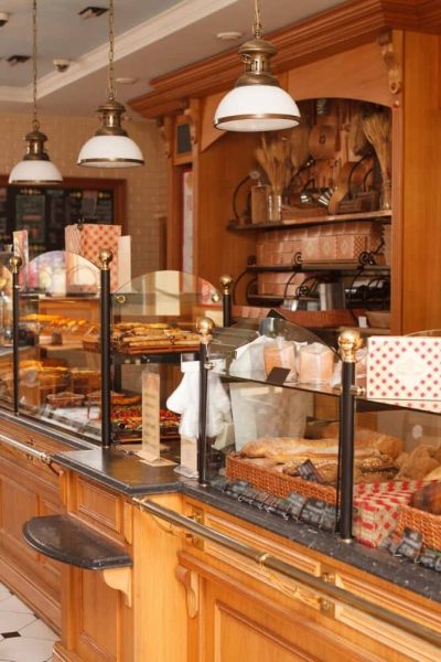 Intérieur d'une boulangerie avec des comptoirs en bois présentant divers pains et pâtisseries derrière une vitre, avec des lampes suspendues et des étagères décoratives en arrière-plan.