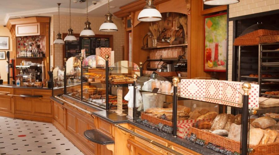 Intérieur d'une boulangerie avec des comptoirs en bois présentant divers pains et pâtisseries derrière une vitre, avec des lampes suspendues et des étagères décoratives en arrière-plan.