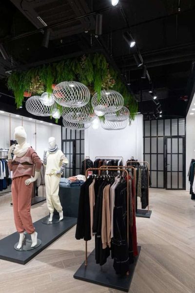 Intérieur d'un magasin de vêtements moderne avec des mannequins présentant des tenues, des étagères de vêtements de part et d'autre, et des plafonniers décoratifs avec de la verdure au-dessus de la tête.