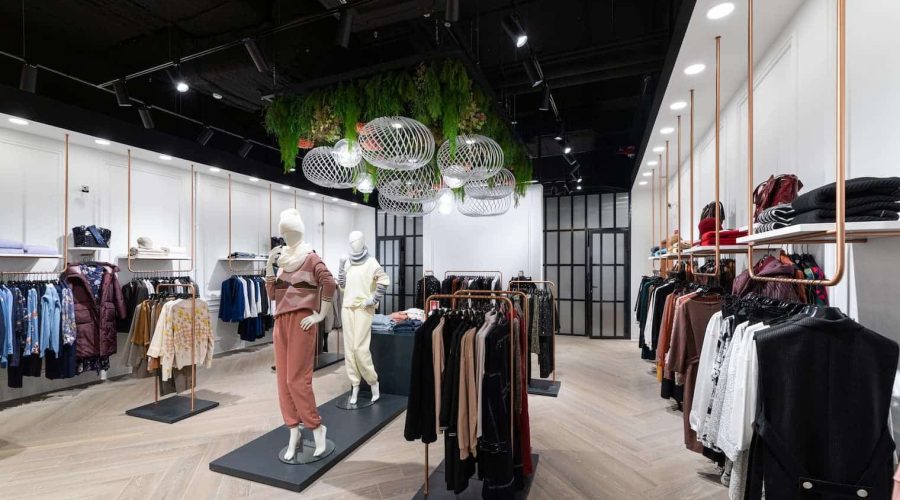 Intérieur d'un magasin de vêtements moderne avec des mannequins présentant des tenues, des étagères de vêtements de part et d'autre, et des plafonniers décoratifs avec de la verdure au-dessus de la tête.
