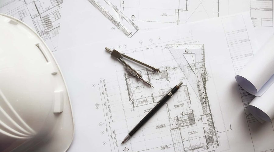 Des plans d'architecture sur un bureau avec un casque blanc, un crayon, un compas, une règle et des plans enroulés.