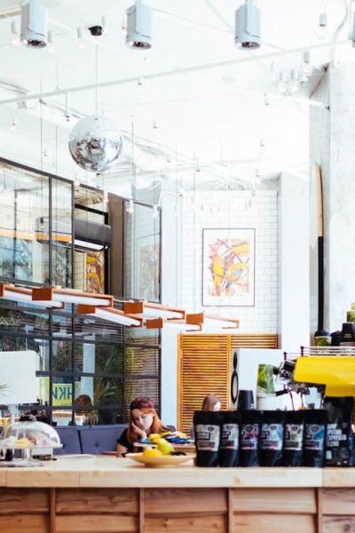 Un intérieur de café moderne et lumineux avec de grandes fenêtres, des plantes, un comptoir en bois présentant des produits de café et une personne assise à une table à l'arrière-plan.