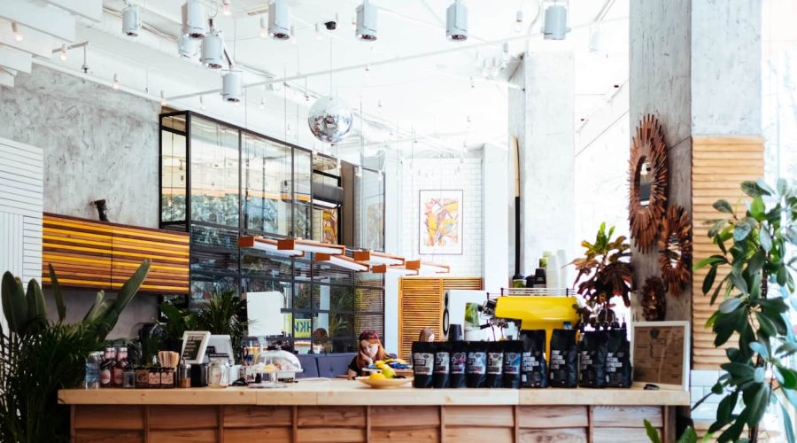 Un intérieur de café moderne et lumineux avec de grandes fenêtres, des plantes, un comptoir en bois présentant des produits de café et une personne assise à une table à l'arrière-plan.