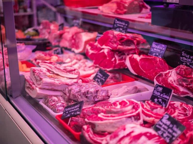 Dans la vitrine d'une boucherie, différents morceaux de viande rouge crue sont exposés, avec de petites étiquettes noires indiquant les types de viande, disposés avec la précision d'un agencement de bureau.