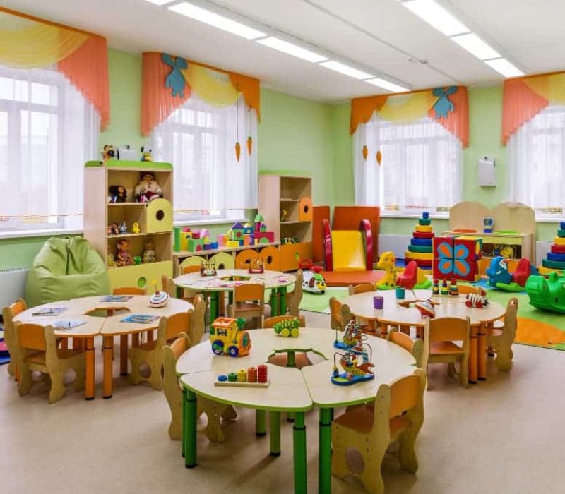 Une salle de classe bien éclairée, équipée de petites tables et de chaises, présente un agencement de bureau bien pensé, complété par des étagères de jouets, des tapis de jeu colorés et des équipements de jeu éducatifs soigneusement disposés dans la pièce.