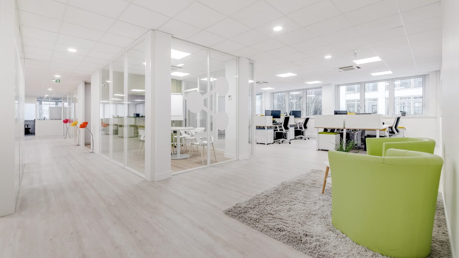 Espace de bureau moderne avec des salles de réunion en verre, des bureaux blancs, des chaises vertes et un tapis gris sur un sol en bois clair, avec de grandes fenêtres apportant de la lumière naturelle.