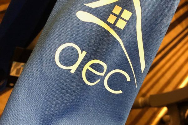 Tissu bleu portant un logo blanc avec une maison stylisée et les lettres minuscules "aec" en dessous.