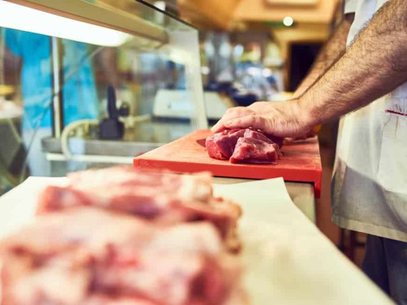 Boucher tranchant de la viande crue sur une planche à découper rouge, avec d'autres morceaux de viande au premier plan sur du papier blanc ; l'espace de travail organisé reflète l'agencement de bureau d'un expert.