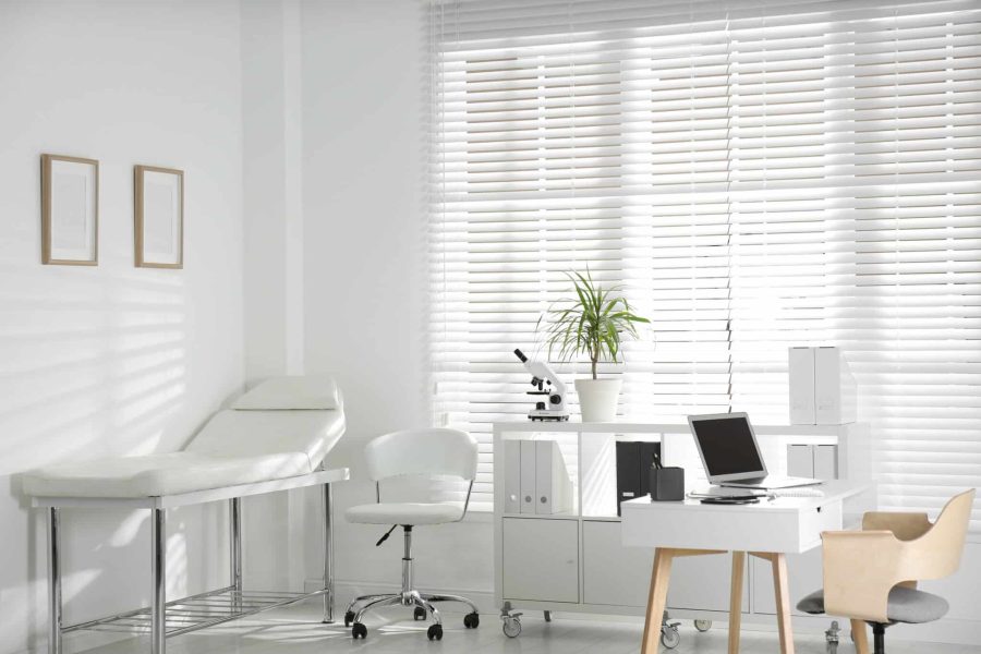 Cabinet médical lumineux équipé d'une table d'examen, d'un bureau, de chaises blanches, d'un ordinateur portable, d'un microscope, d'une plante en pot et de grandes fenêtres dotées de stores.