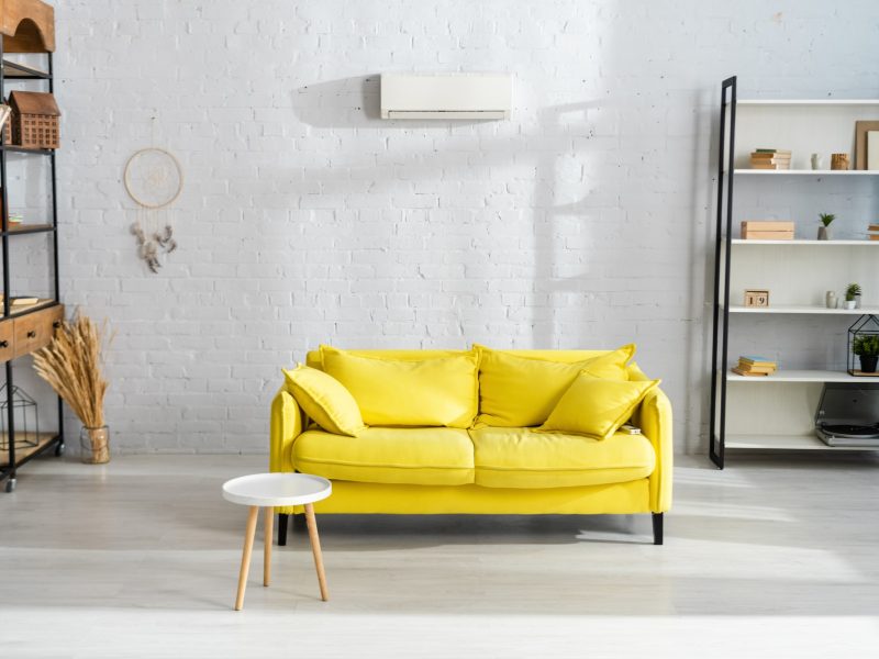 Un canapé jaune vif, agrémenté de coussins, trône au centre d'un salon moderne aux murs blancs, avec des étagères et une petite table d'appoint ronde blanche.