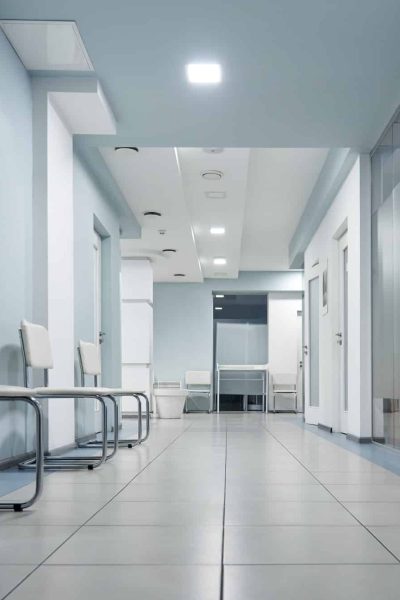 Couloir vide dans un bâtiment médical ou administratif avec des chaises blanches alignées contre le mur, un sol carrelé et des portes fermées des deux côtés.