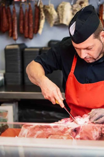 Un boucher en tablier orange et casquette noire découpe de la viande crue sur un comptoir, avec des saucisses suspendues et une trancheuse à viande visibles à l'arrière-plan.
