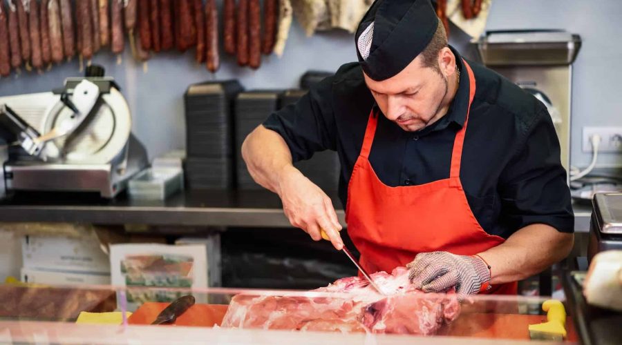 Un boucher en tablier orange et casquette noire découpe de la viande crue sur un comptoir, avec des saucisses suspendues et une trancheuse à viande visibles à l'arrière-plan.