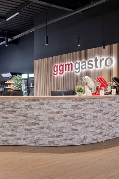 Un comptoir d'accueil moderne avec une façade en briques chez GGM Gastro, présentant des plantes, des articles d'exposition et des réfrigérateurs sur un fond noir et boisé.