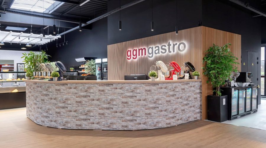 Un comptoir d'accueil moderne avec une façade en briques chez GGM Gastro, présentant des plantes, des articles d'exposition et des réfrigérateurs sur un fond noir et boisé.
