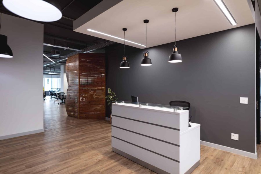 Espace d'accueil moderne dans un bureau, avec un bureau blanc, une chaise noire, des suspensions lumineuses, un parquet et un mur incurvé en bois en arrière-plan.