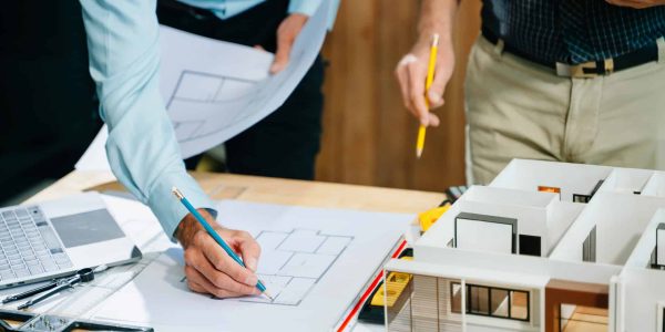 Deux personnes examinent et annotent des plans d'architecture sur un bureau où se trouvent également un ordinateur portable, une règle graduée et une maquette.