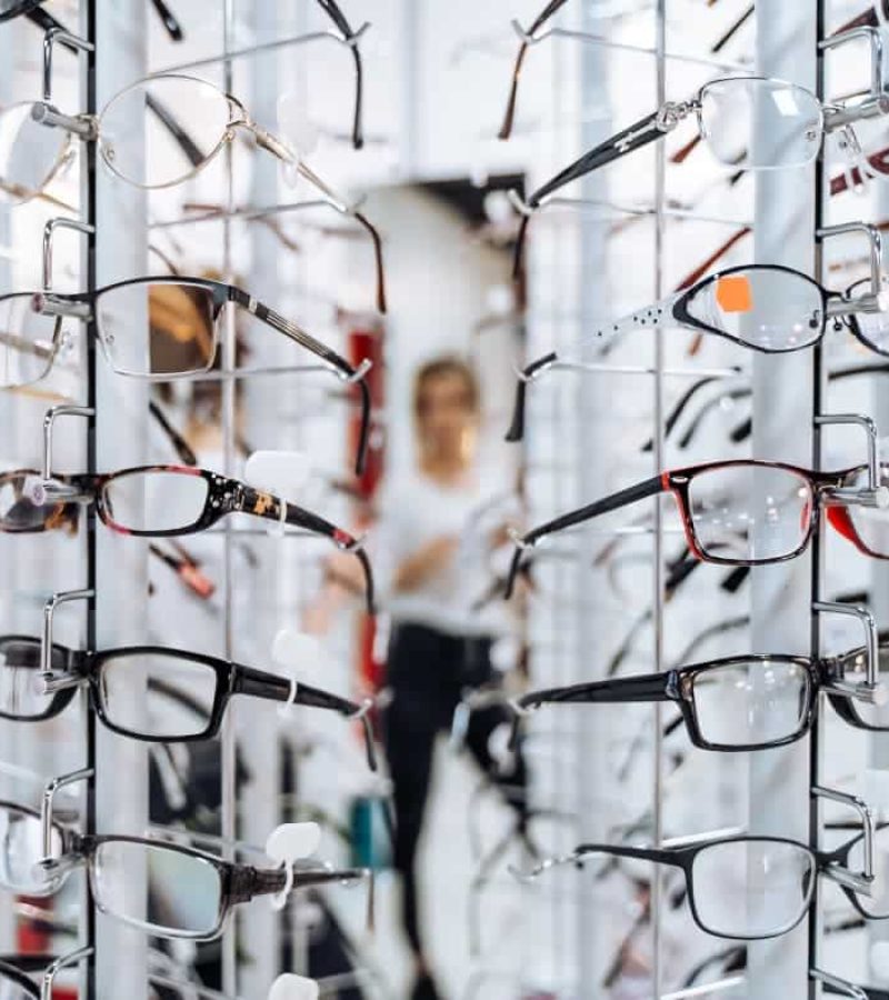 Présentoir de lunettes de différents styles et couleurs ; une personne apparaît en arrière-plan, mettant en évidence l'agencement moderne d'un bureau à l'intérieur d'un magasin de lunettes.