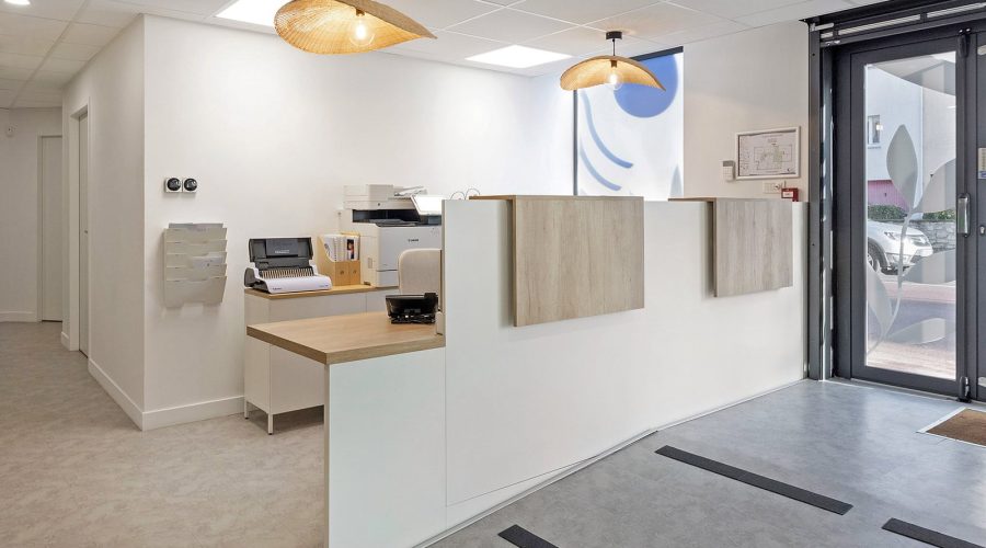Espace de réception moderne avec un bureau blanc, des accents en bois, des équipements de bureau, des lampes suspendues et des portes d'entrée en verre.