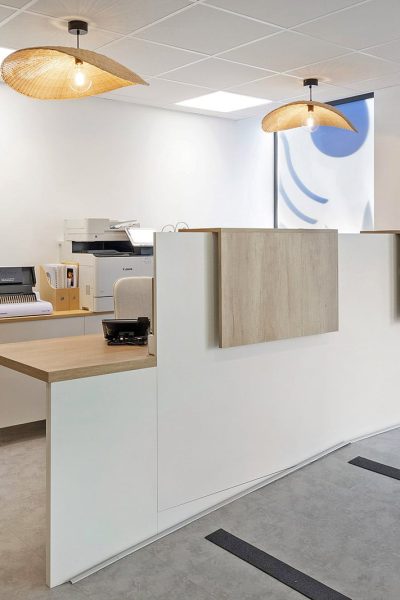 Espace de réception moderne avec un bureau blanc, des accents en bois, des équipements de bureau, des plafonniers et une porte d'entrée en verre.