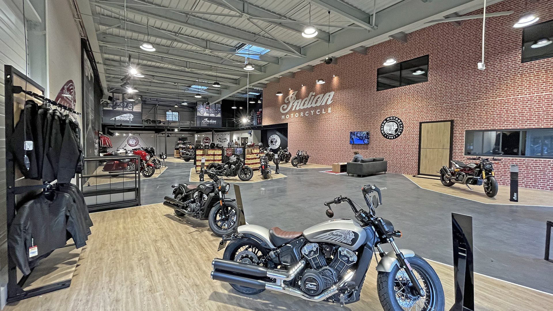 Salle d'exposition de motos spacieuse avec plusieurs motos en exposition, un mur d'accent en briques avec le logo Indian Motorcycle et des racks d'équipement de moto sur le côté.