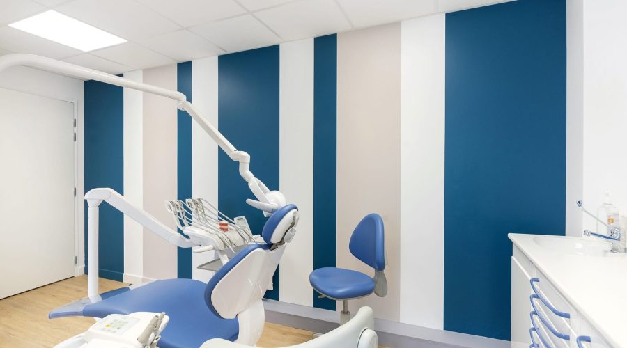 Salle de soins dentaires moderne avec murs rayés bleu et blanc, fauteuil dentaire, équipement et tiroirs de rangement ; intérieur clair et propre.