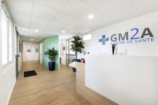 Réception d'un centre médical avec un bureau blanc, des plantes d'intérieur, un sol en bois clair et un panneau indiquant "GM2A Centre de Santé" sur le mur.
