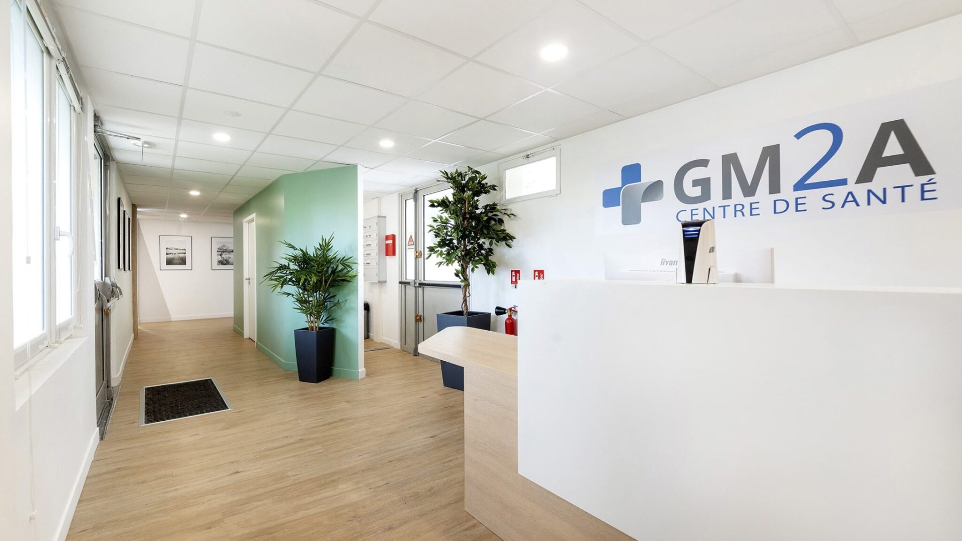 Réception d'une clinique médicale moderne avec un bureau blanc, des plantes vertes, un sol en bois et l'inscription "GM2A CENTRE DE SANTÉ" sur le mur.