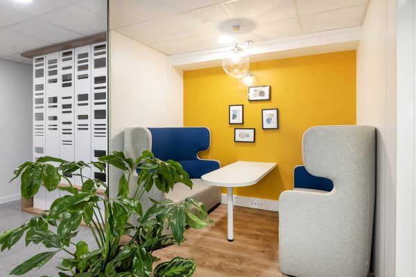 Petit coin de réunion de bureau avec deux cabines grises et bleues, une table blanche, un mur d'accent jaune, des photos encadrées et une plante au premier plan.