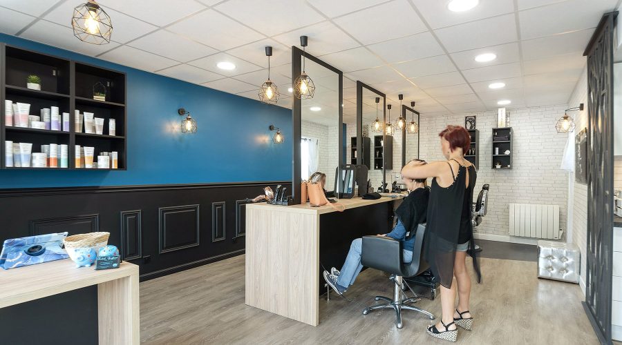 Une coiffeuse coiffe une cliente dans un salon moderne aux murs bleus et blancs, au sol en bois et aux étagères présentant des produits capillaires.