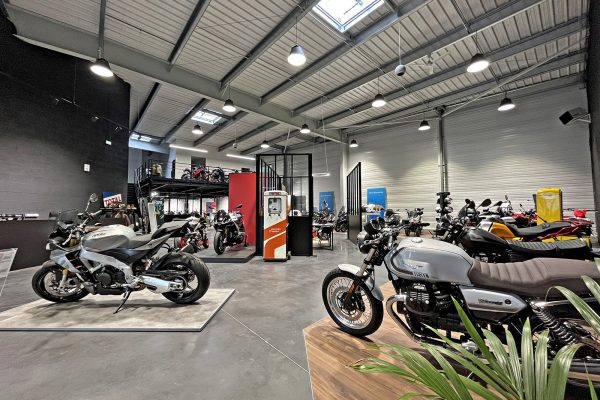 Intérieur d'une salle d'exposition de motos moderne avec plusieurs motos exposées, de hauts plafonds, un éclairage industriel et une mezzanine à l'arrière-plan.