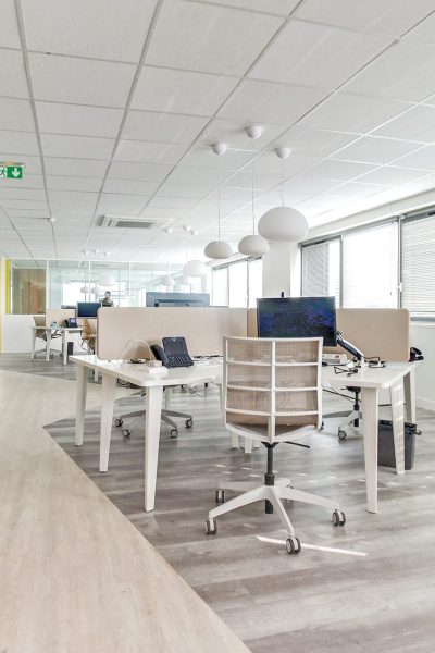 Espace de bureau moderne avec des bureaux blancs, des chaises, des ordinateurs et de grandes fenêtres ; l'espace est propre, lumineux et décoré de façon minimale.