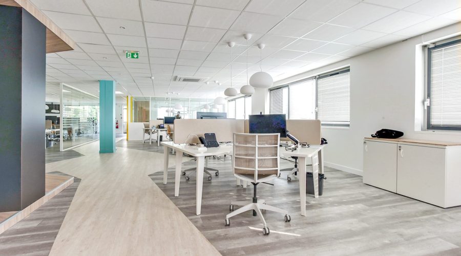 Espace de bureau moderne avec des bureaux blancs, des chaises, des ordinateurs et de grandes fenêtres ; l'espace est propre, lumineux et décoré de façon minimale.