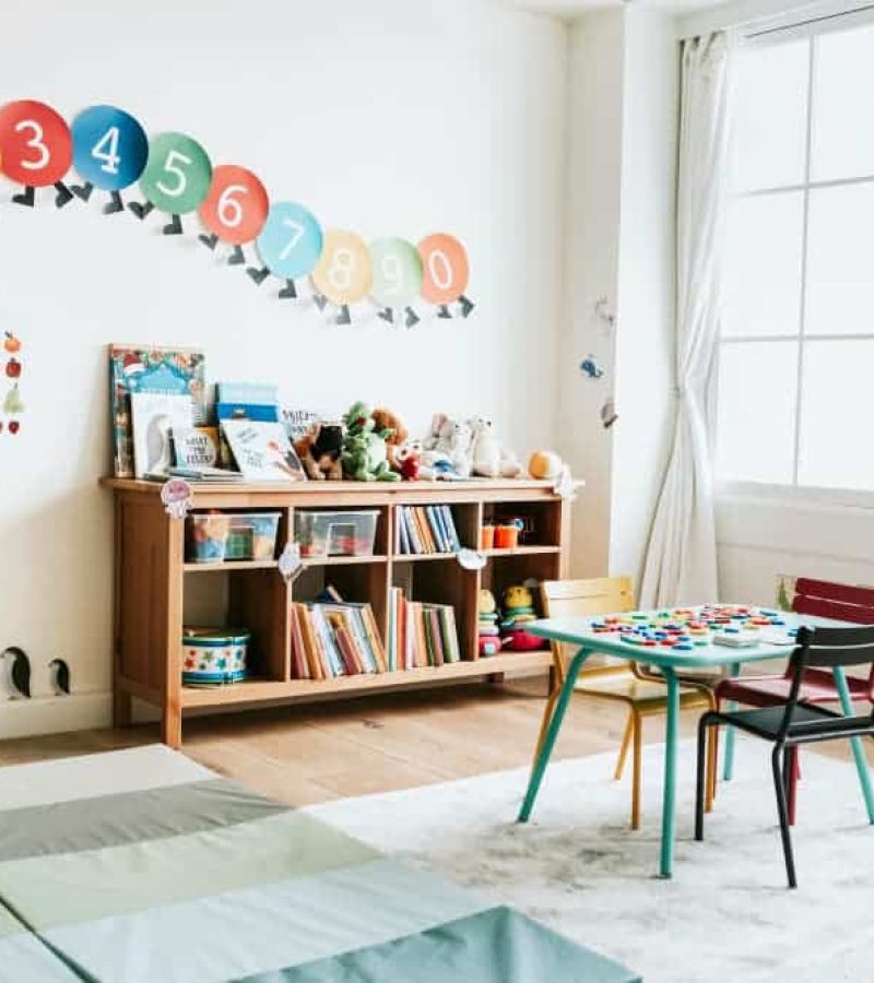 Une salle de classe lumineuse avec une étagère, une table et des chaises pour enfants, des tapis et des décorations colorées de chiffres et de fruits sur les murs blancs est un exemple d'agencement de bureau réfléchi pour les jeunes apprenants.