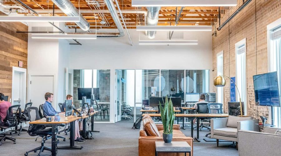 Bureau moderne à aire ouverte avec des personnes travaillant à des bureaux, un plafond apparent, une salle de réunion en verre, un coin salon avec canapé et de grandes fenêtres laissant entrer la lumière naturelle.