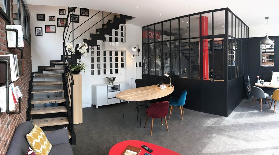 Bureau moderne avec un escalier, une salle de réunion vitrée, une table ronde avec des chaises colorées et des œuvres d'art sur un mur blanc et brique.